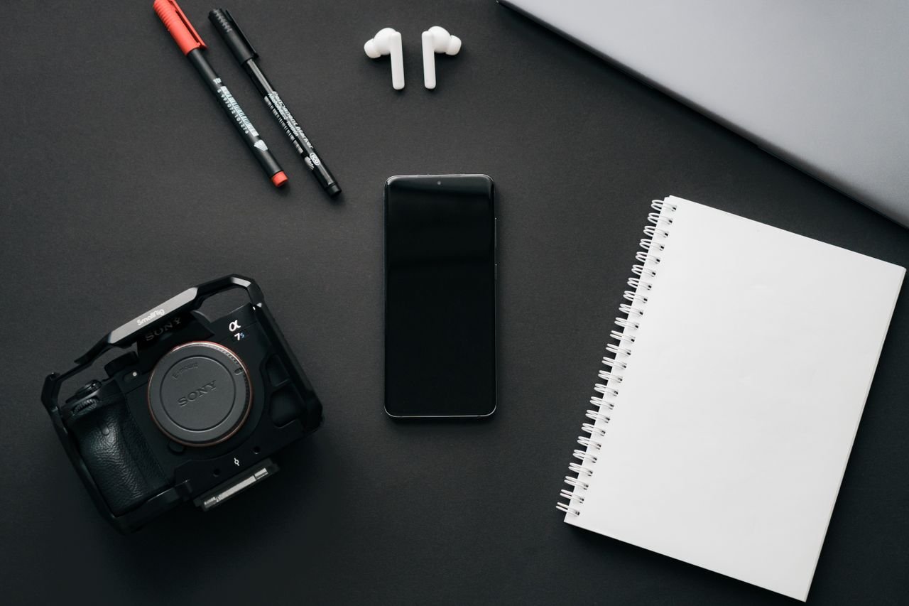 services-03 Flatlay of modern gadgets including a camera, smartphone, and notebook on a black surface.