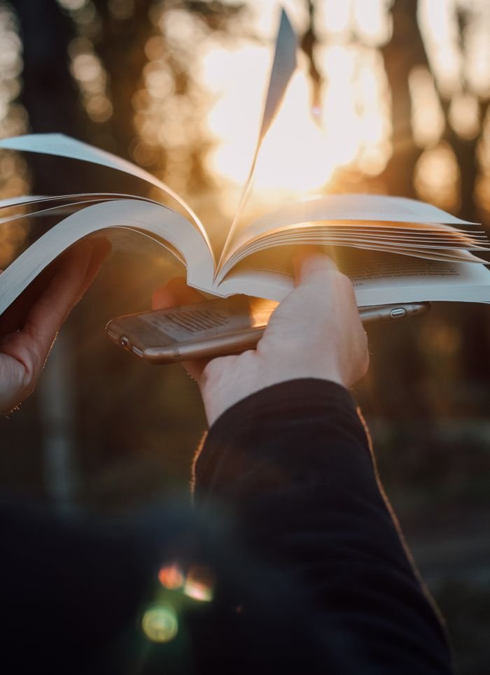 Close-up of hands holding a book and smartphone with sunlight filtering through the trees.