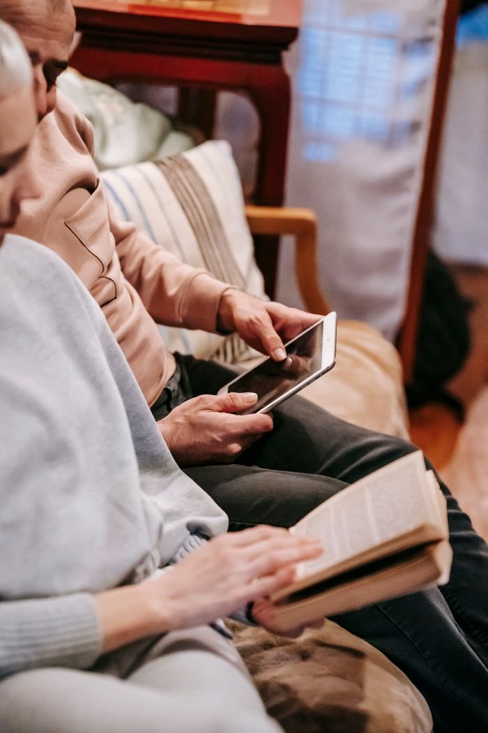 Crop unrecognizable couple reading book and using tablet while relaxing in comfortable sofa at home